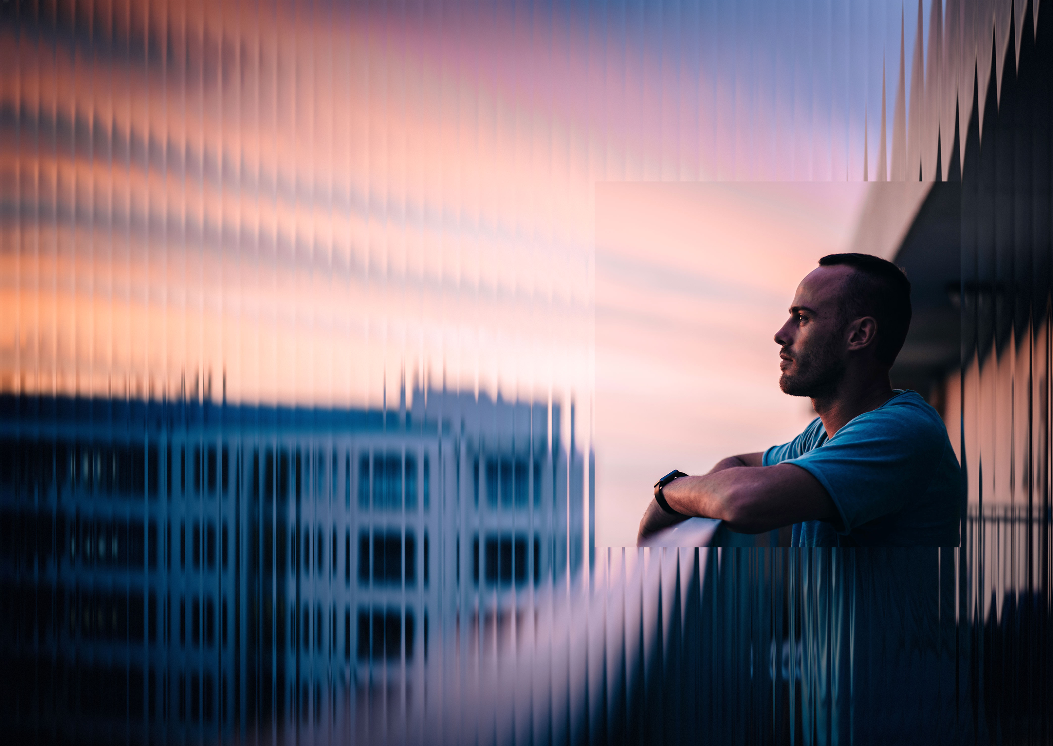 A man in a teal shirt looking out from a balcony at dusk, seen through glass.